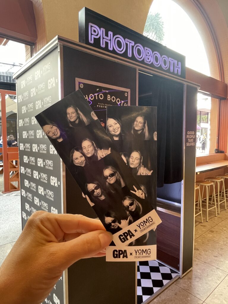 Hand holding two branded photo strips in front of a retro enclosed photo booth at a Gold Coast venue permanent install